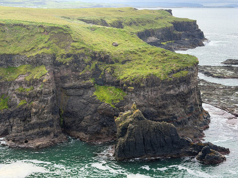 Rugged basalt cliffs topped with green grass rise above a dark volcanic sea stack and Atlantic waters along the Giant&rsquo;s Causeway coast in Northern Ireland.