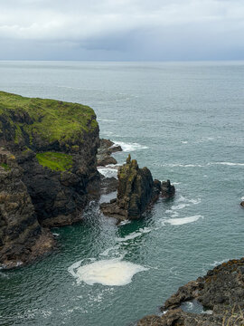 A jagged basalt sea stack rises from Atlantic waters below grass-covered volcanic cliffs along the Giant&rsquo;s Causeway coast in Northern Ireland.