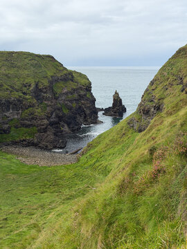 A rugged basalt sea stack rises from a narrow rocky inlet framed by steep green cliffs along Northern Ireland&rsquo;s Giant&rsquo;s Causeway coast.