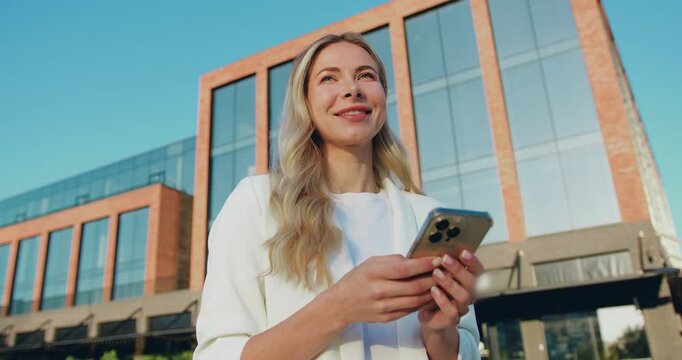 Young businesswoman standing on city street, using her smartphone to send a text message, smiling and looking away.