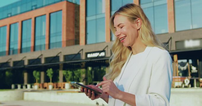 Side view of an adult blonde woman with long hair holding a smartphone, scrolling through online pages and smiling while standing on a city street.