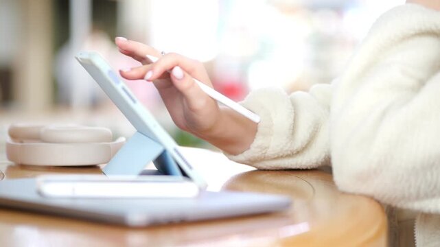 Woman in cozy sweater uses tablet with stylus. Hands focus on screen. Warm wooden table surface. Soft indoor lighting. Blurred background shows cafe-like setting.