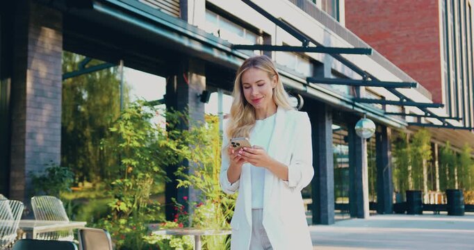 Elegant businesswoman wearing a white blazer walking along a city street, using her smartphone and smiling.