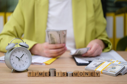 In a lively office environment, a person examines stacks of cash while holding bills. An alarm clock and coins symbolize the value of time in financial decisions.