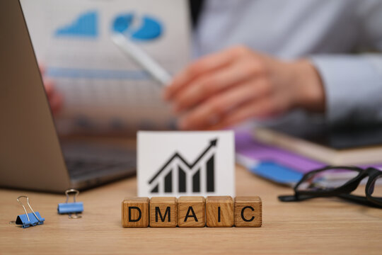 A person studies charts and graphs while seated at a workspace. Blocks spelling DMAIC sit on the table, symbolizing a structured approach to enhancing operational performance.