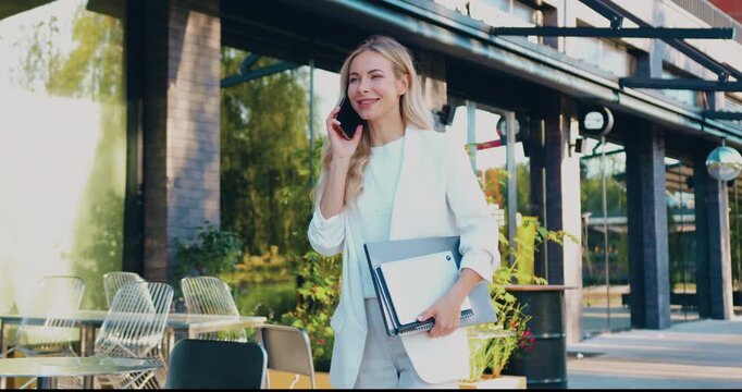 Young woman in white suit walking through city while talking on phone and carrying laptop and notebook