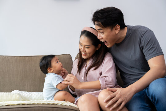 Asian parents gently hold and interact with their baby's hands while sitting together on a sofa, illustrating close parental care, affectionate bonding, family connection, and everyday home life.