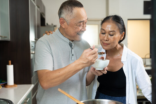 Senior Asian couple share a warm moment while preparing food in their home kitchen, showing affection, companionship, everyday cooking, and a peaceful lifestyle together.