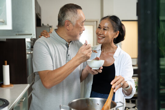 Senior Asian couple share food and affection in a home kitchen, with the man offering a spoonful while the woman holds a bowl, illustrating companionship and everyday partnership.