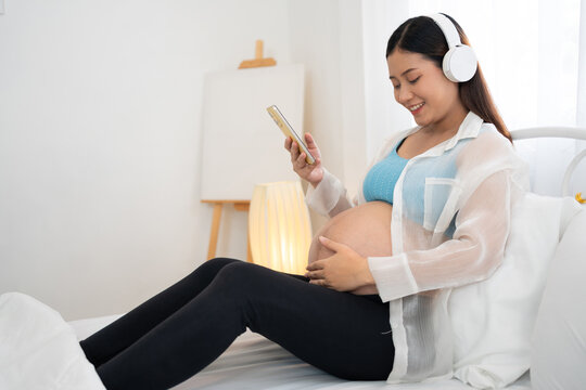 Pregnant woman relaxes on a bed while using a smartphone and wearing headphones, combining maternity lifestyle, digital communication, music listening, and comfortable home living.