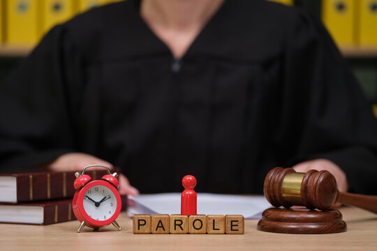 A judge in a black robe sits at a wooden desk surrounded by law books, an alarm clock, and a gavel. A red figure signifies a parole decision, initiating a serious discussion about freedom.