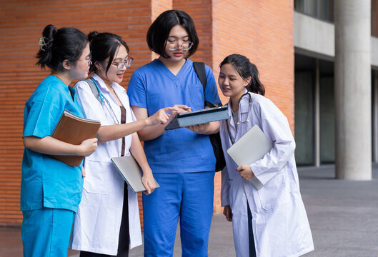 Asian healthcare students in white coats and scrubs discuss coursework together outside a university building, using tablets and notebooks in a collaborative campus learning setting.