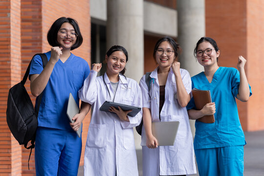 Asian healthcare students celebrate together outside a university building, raising their fists with smiles while carrying study materials and technology during an energetic campus moment.