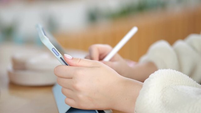 Hands use smartphone and stylus on tablet. Soft focus on cozy indoor setting. Person wears white fleece sleeve. Wood table surface visible. Blurred background with warm tones
