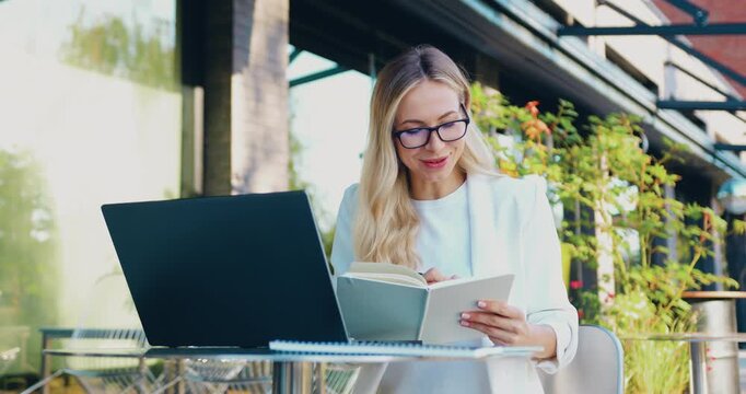 Woman taking notes in a notebook while watching an online video lesson on a tablet at a table on a busy urban street, studying remotely through digital education technology.