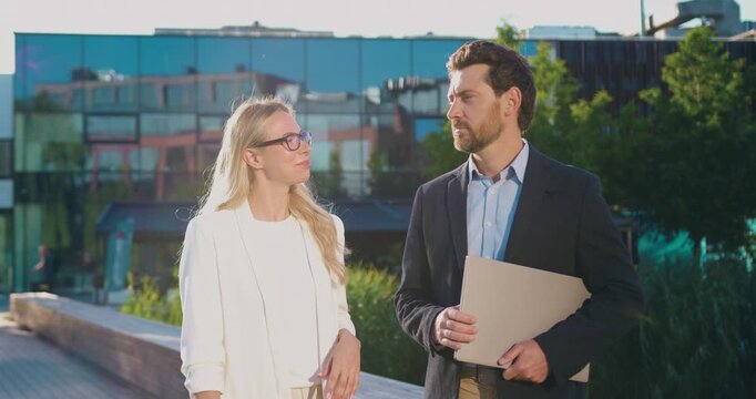 Young professional couple man and woman walking outdoors along modern glass office buildings holding laptop during a sunny afternoon with engaged friendly conversation sidewalk