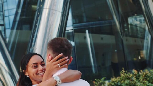 A young couple embracing and smiling affectionately in an outdoor urban setting with modern glass architecture and greenery during daytime.