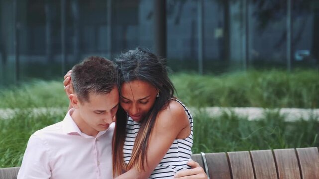 A mixed-race couple embraces affectionately while sitting on a wooden bench in a lush urban park during daytime. They share a tender moment surrounded by greenery and modern buildings.