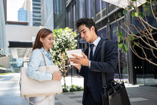 Professional business colleagues reviewing project data together on a digital tablet outdoors while standing outside office buildings during a workday