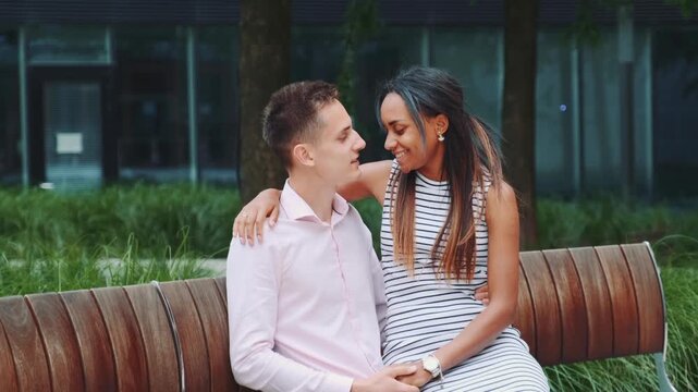 A light-skinned man and dark-skinned woman share an intimate moment sitting closely on a bench in a green urban park during daytime.
