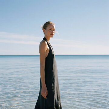 Woman in black halter dress standing in calm shallow sea background for fashion banner and summer lookbook and editorial campaign