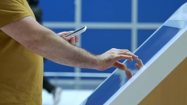 Man uses touch screen kiosk with phone in hand. Blue background, indoor setting. Person interacts with digital display. Hand gestures shown on glass surface. Modern technology interaction scene