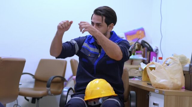 Worker in uniform wearing safety helmet and hard hat makes hand gesture beside table equipment during indoor workshop maintenance near chair, desk, tools, and protective gear