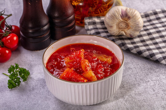 Bowl of vibrant red lecho with chunky peppers and tomatoes, surrounded by garlic, cherry tomatoes, parsley, and wooden spice grinders on textured gray surface