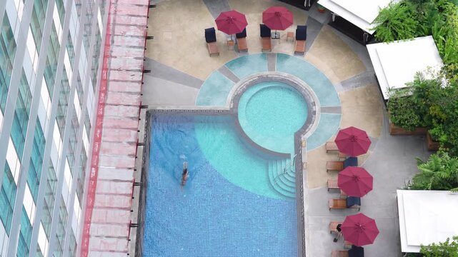 Aerial View of Rooftop Swimming Pool at Urban Hotel Terrace with Turquoise Water, Red Umbrella, Lounge Chairs, Circular Spa, Swimmer, Tropical Courtyard