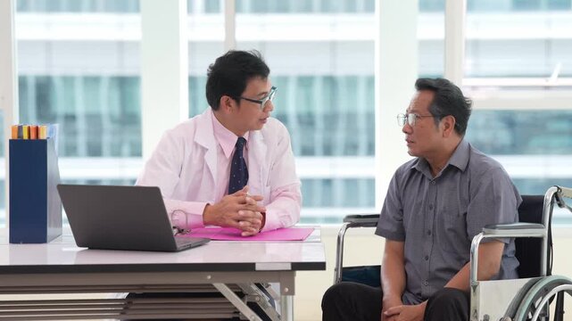Doctor consultation with patient in wheelchair at healthcare clinic office, discussing treatment and rehabilitation during appointment beside laptop, documents, workplace paperwork and supportive care