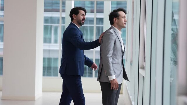 Businessman in gray suit at office window with colleague during business meeting, professional executive viewing modern corporate interior and building facade, thoughtful workday posture