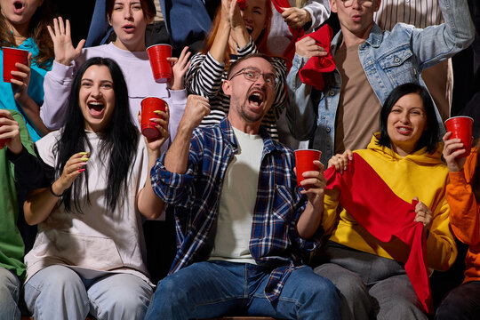 Cheering volleyball supporters raise hands and drinks in arena seats with contagious joy. Sports fandom, game night and group celebration concept.