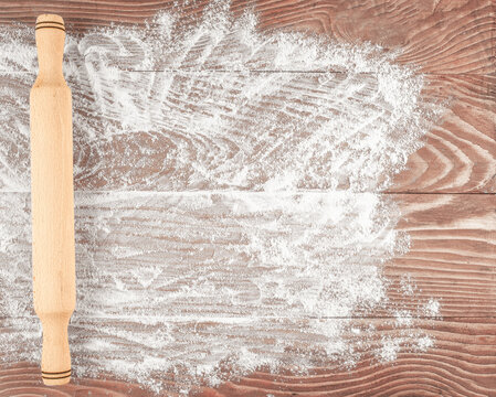 Wooden rolling pin beside scattered flour and circular hand drawn marks on rustic brown plank surface, overhead baking preparation scene with textured kitchen workspace. copy space