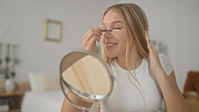 Woman applying mascara to eyelashes with a wand while holding a mirror in a bedroom; self care calm.