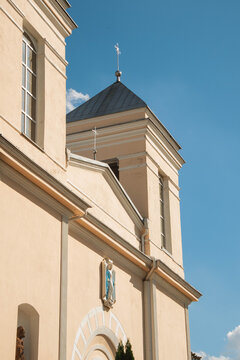 Catholic church facade and bell tower viewed from below, featuring beige walls, classical architectural details, arched niches, a religious statue, a steep roof with cross, and a clear blue sky