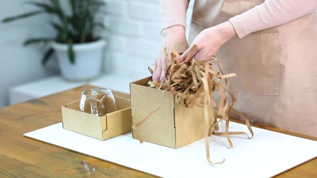 Woman putting crinkle paper filler into cardboard box while packing fragile item. Small business order packaging and gift box shipping preparation concept