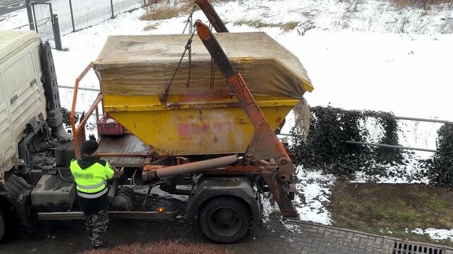 A worker in a high visibility jacket operates a truck with a hydraulic lift to load a covered yellow waste container, demonstrating industrial waste management and logistics tasks