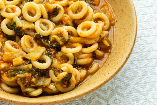 Close-up of an authentic Italian Anelli Siciliani con Bietole e Pomodori dish. A brown ceramic plate with ring-shaped pasta on a bright background featuring a beige geometric pattern.