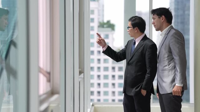 Businessman and colleague in suit discuss work beside office window during professional business meeting, pointing toward urban building view in modern corporate workplace conversation