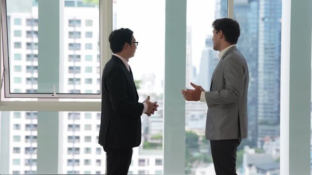 Businessman and male colleague in suit lead business meeting discussion beside office window, professional corporate conversation on urban planning, city skyscraper views and partnership