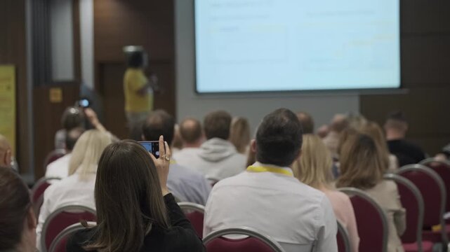 Audience watching speaker and slides during business conference. Woman records presentation on smartphone. Professional seminar setting, networking, learning, engagement.