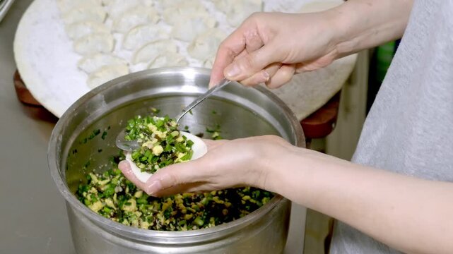 A person prepares homemade dumplings, holding a metal bowl of vegetable and egg filling, with shaped dumplings and wrappers nearby.