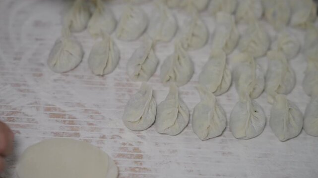 A person prepares uncooked dumplings, holding one, with stacked wrappers and finished dumplings on a floured surface.