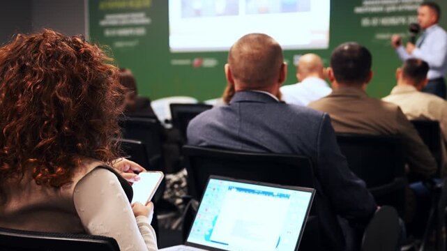 People attend a professional conference, listening to a speaker and recording important information with digital devices during a presentation.