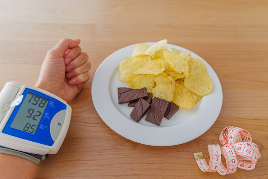 Blood pressure monitor displaying high reading next to plate with potato chips, chocolate, and measuring tape on wooden table, unhealthy lifestyle and hypertension risk concept
