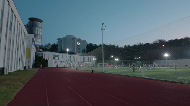 An outdoor sports complex with red running track and grass football pitch, lit at dusk with surrounding buildings.