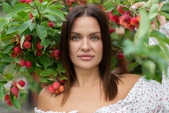 Smiling young woman in a white floral dress surrounded by red crabapples and green leaves in a fruit orchard. Natural outdoor portrait with a joyful summer mood