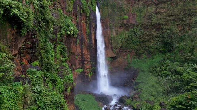 Aerial view of Kapas Biru Waterfall surrounded by lush tropical rainforest in East Java, Indonesia. Scenic jungle landscape with cascading water, mist and dense green vegetation.