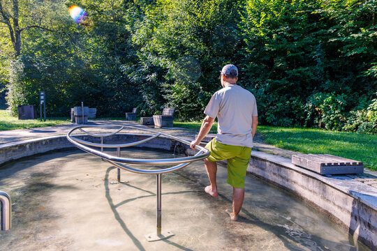 Man walking barefoot in outdoor Kneipp water treading pool surrounded by green trees on sunny day, hydrotherapy wellness and active lifestyle concept