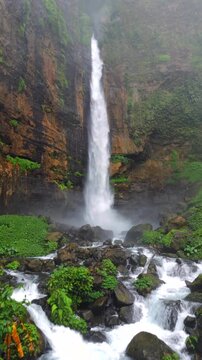 Kapas Biru Waterfall with a rocky stream flowing through lush tropical rainforest in East Java, Indonesia. Misty jungle landscape surrounded by cliffs and dense vegetation.